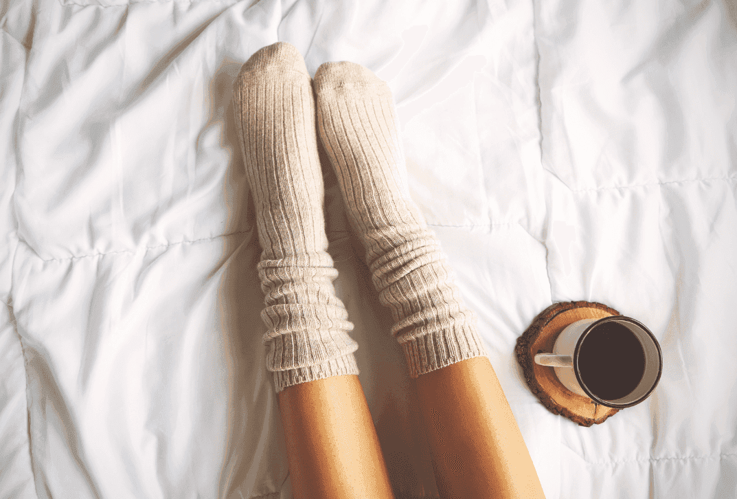 Pale legs with beige wool socks above the ankle laying in bed. Coffee cup on a coaster next do the legs, also on the bed. Bedspread is white crinkled top sheet.